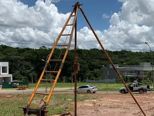 Tripé de sondagem montado num lote de terra, com o martelo suspenso pela haste e o guincho preso à escada.