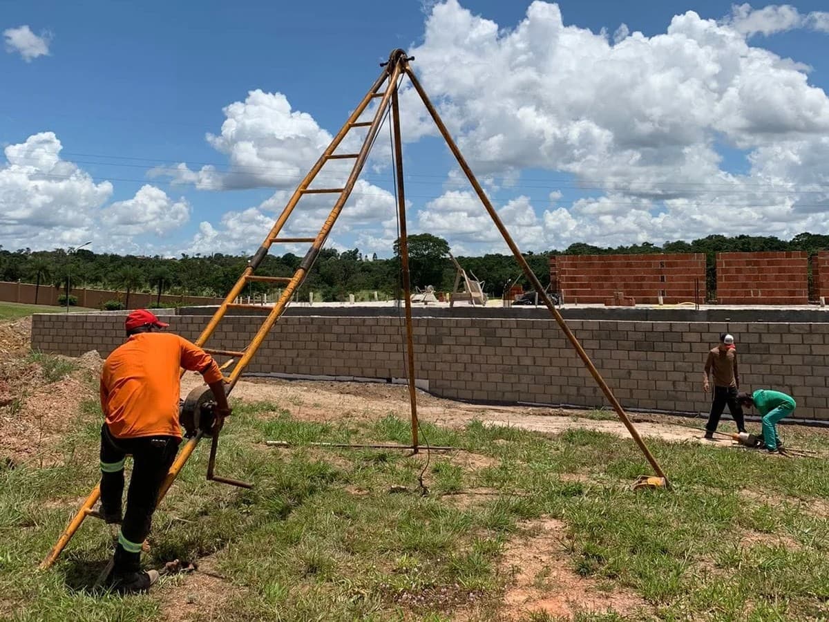 Operador no guincho do tripé de sondagem, num lote cercado por muro de blocos, com a equipe preparando o material ao fundo.